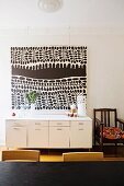 Modern black and white painting above white sideboard next to simple, wooden armchair with cushion