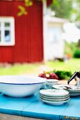 Stack of plates and enamel bowl on blue-painted garden table with Swedish, Falu red wooden house in background