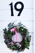 Aufgehängter Weihnachtskranz aus Tannenzweigen und Schleifenband an weisser Holztür unter Hausnummer