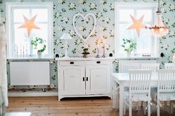 Dining room with floral wallpaper, white-painted, country-house-style furniture and festive star ornaments in windows