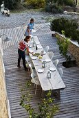 Blick von oben auf lange Tafel aus verwittertem Holz und weiss Stühle, auf Holz Terrassenboden vor mediterranem Garten, Frauen beim Tisch decken