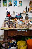 Paints and paintbrushes on vintage work table below postcards of artworks pinned to wall of painter's studio