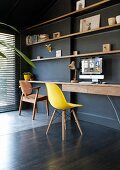 Classic yellow shell chair at minimalist desk below wooden shelves on black-painted wall