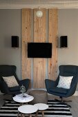 Side tables and swivel armchairs in front of flatscreen TV on wooden panels on wall flanked by loudspeakers