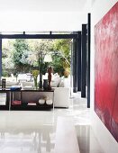 Glossy, white tiled floor in modern interior with open sideboard behind sofa opposite open, folding terrace doors with view into garden