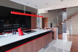 Long kitchen counter with wooden base cabinets below pendant lamp with red-painted narrow housing in modern kitchen with staircase in background