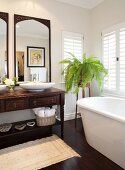 Bathtub below window, antique washstand with countertop basins below framed mirrors and fern on plant stand in corner