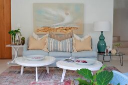 Two coffee tables with white, amorphous tops and pale blue sofa below landscape on wall