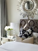 Scatter cushions on bed against upholstered headboard with ornate pattern below mirror with sunburst frame