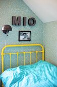 Yellow, vintage metal bed frame, pale blue and white polka-dot bed linen, charcoal wall lamp and decorative letters on patterned wallpaper in corner of teenager's bedroom