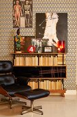 Classic Eames Lounge Chair with black leather cover and matching footstool in front of bookcase and artworks on wall with retro, graphic-patterned wallpaper