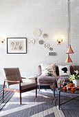 Fifties armchair and grey sofa around coffee table below pendant lamps with copper-coloured lampshades in loft-style interior with decorative wall plates on whitewashed brick wall