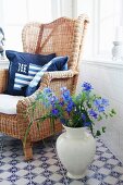 Blue cushion on wicker chair and vase of delphiniums on blue and white, patterned, tiled floor
