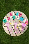 Besteck aus Plastik und Holz in Pastelltönen und mit bunten Buchstaben gelegter schwedischer Text auf Gartentisch in Rasenfläche