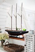 Splendid model of sailing boat on half-height wall in Scandinavian interior; wicker tray and vintage lantern in foreground