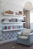 Reading corner with pale armchair next white bookshelves mounted on wallpaper with geometric pattern