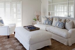 Polsterhocker mit weisser Husse und passendes Sofa mit drapierten Kissen in elegantem Wohnraum mit maritimem Flair