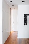 Hallway with white fitted cupboards and wooden floor; black jacket hanging from coat rack on wall