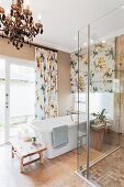 Floral gemusterte Vorhänge und ein Kronleuchter als Kontrast zu Designerbadewanne und moderner Duschabtrennung aus Glas