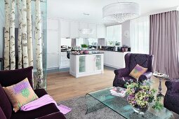 A view from a seating area with purple upholstered furniture and an acrylic table looking towards a white, fitted kitchen with an island counter