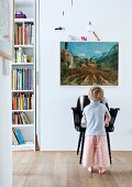 Young child in front of black armchair below landscape painting on wall