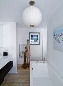 Modern bathtub below white, spherical lamps, washstand to one side and unfinished branch used as clothes rack in background