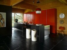 Cylindrical bar stools with footrests on slate floor at black, free-standing kitchen island in front of red fitted cupboards on wooden wall in open-plan interior