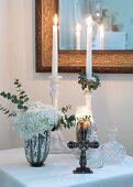 Brennende Kerzen auf Kerzenständer und religiöses Kreuz vor Kristallglas Flakons auf Tisch mit weißem Tischtuch