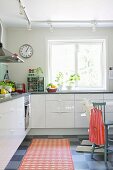 Simple, white fitted kitchen with orange rug on chequered floor