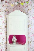 White, rustic corner cabinet with carved pediment against vintage-style floral wallpaper