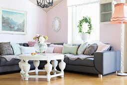 Round, white coffee table with carved base in front of corner sofa against pastel pink wall