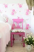 Pink-painted chairs with carved backrest and seat cushions against floral wallpaper next to pot of flowers on floor