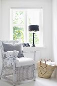 Comfortable, white wicker chair with seat cushion and raffia shopping bag with heart motif in front of black table lamp on windowsill in simple interior