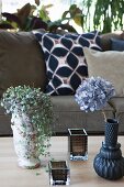 Foliage plant in vintage plant pot, hydrangea in vase and tealight holders on table in front of sofa with scatter cushions
