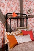 Orange retro standard lamp next to black metal bed with collection of scatter cushions in shades of orange and wallpaper with pattern of coral
