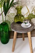 Green carafe and drinking glass on vintage wooden stool next to emerald-green floor vase against wall with leaf-patterned wallpaper