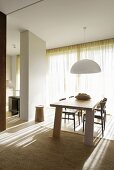 Pendant lamp with white lampshade above pale wood dining table and chairs in front of sunlit glass wall with closed curtain; partition screening kitchen to one side