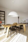Pale, solid wooden table and dark wooden chairs below pendant lamp with white lampshade in minimalist interior; pattern of light and shade on floor in foreground
