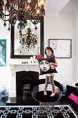 Little girl holding cushion standing on classic chair next to open fireplace below mirror and chandelier in traditional interior with black and white accents