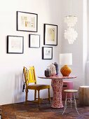 Vases and table lamp on table covered in floral wallpaper and various chairs below framed pictures on white wall
