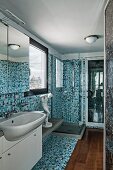 Narrow bathroom with mosaic tiles in various shades of blue, white washstand with mirrored cabinet and wooden floor section
