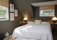 Dunkelgrau getöntes Schlafzimmer im Dachgeschoss, Doppelbett mit weisser Tagesdecke, unter kleinem Sprossenfenster, links und rechts Nachtkästchen mit Tischleuchte
