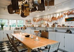 Kupfer Kochgeschirr an Wand über Küchenzeile mit weisslackierten Holzunterschränken, langer Holztisch unter an Decke aufgehängter Korbsammlung