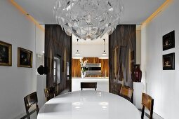 Prunkvolle Kristalllampe über weißem, ovalen Esstisch vor Durchgang zu moderner Küche mit holzfurnierten Fronten