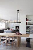 Classic chairs at pale wooden table, collection of white candles on shelf suspended from ceiling, open-plan kitchen area with island counter in background and fitted book shelves to one side