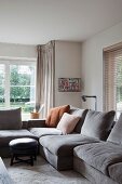 Corner of modern living room - grey velvet sofas, round footstool with cushion on carpet and lattice windows with view of garden in background