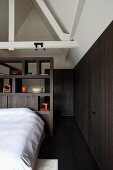Schlafzimmer unter dem ausgebautem Dach - Bett mit weisser Bettwäsche vor dunklem Holzregal als Raumteiler, seitlich dazu passender Einbauschrank aus dunklem Holz