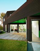 Simple wooden terrace and garden in front of illuminated interior of contemporary house with dark wood cladding