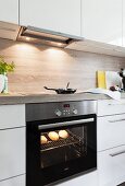 Bread rolls in an illuminated often and a frying pan on a ceramic glass hob in a built in kitchen with white cupboards and a wooden look work surface