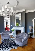 Armchairs with grey velvet upholstery on zebra-skin rug and elegant chandelier in front of mirror above fireplace